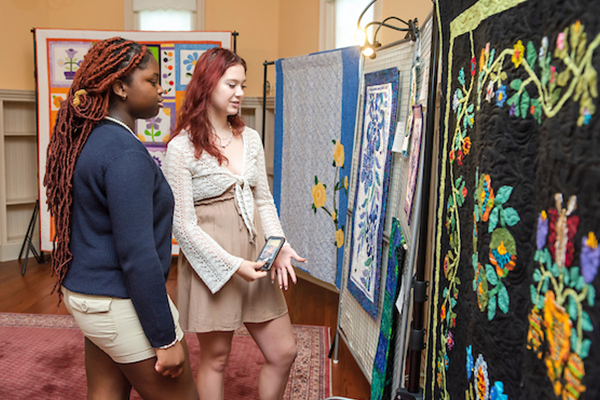 In a yellow room in a historic building, two young women admire a quilted artwork hanging from a metal frame. Behind them, five other quilts are displayed from frames around the perimeter of the room. There is a large oriental rug on the hardwood floor.