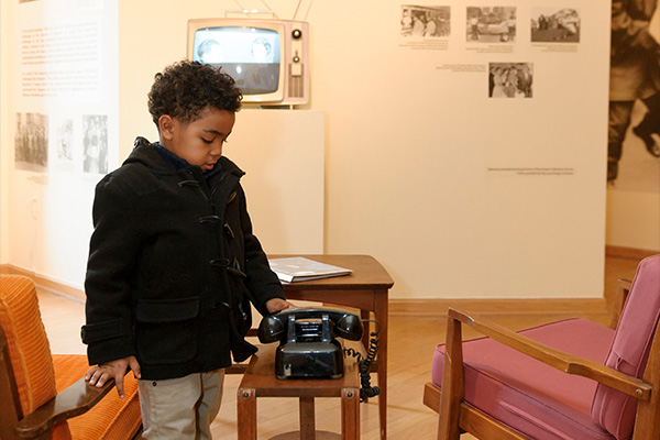 A child in a black coat standing in a interactive set piece of chairs, a table, and an old television. The set is designed to look like a 50s household, and has information regarding civil rights activists in America.