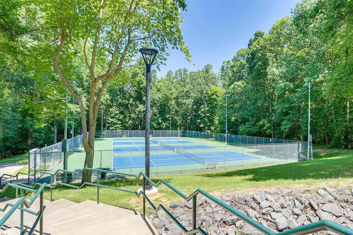 Four sunny tennis courts surrounded by a chain-link fence and tall trees. In the foreground is a concrete staircase leading down past the courts.