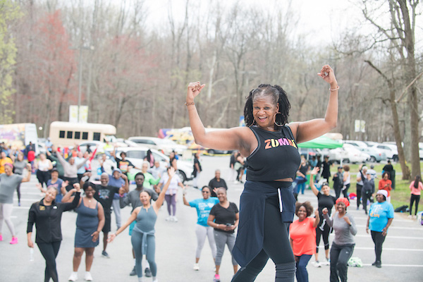 A woman with a shirt that says "Zumba" exercising and demonstrating for a crowd of people behind her.