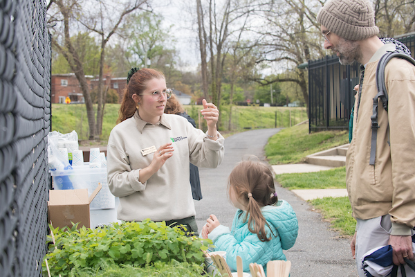A father and child speak with a staff member standing outside near a tray of seedlings.