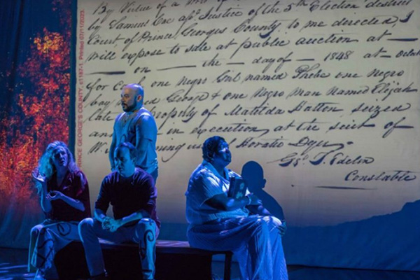 Four performers lit with blue lights on a stage, with a backdrop of a historic, handwritten document referring to a slave auction in Prince George's County.