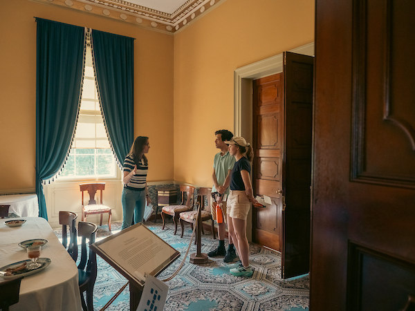 A guide speaks to two visitors in a yellow room in a historic building. Tables and chairs in the room are part of an exhibit behind a rope.