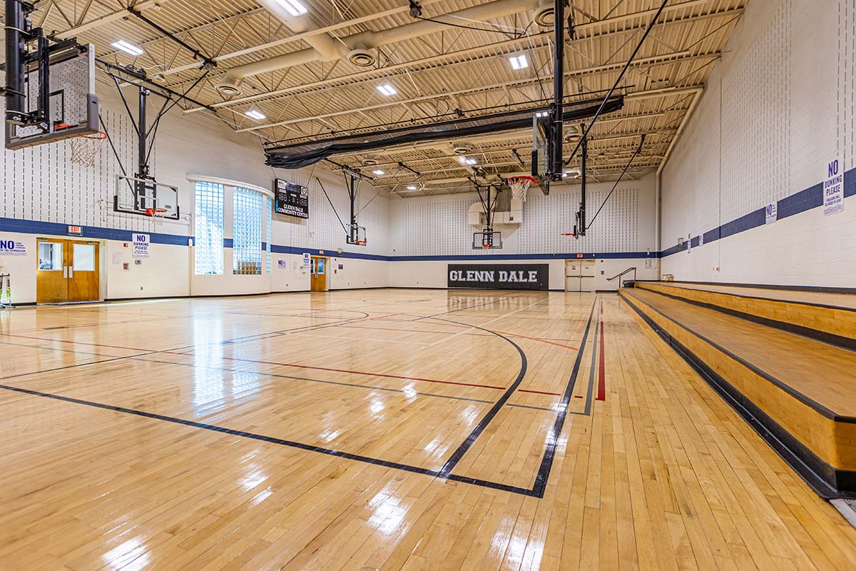 A gymnasium with wooden bleachers down one wall, and a curved, glass-block window in the opposite wall, next to a scoreboard. On the far wall are the words Glenn Dale.
