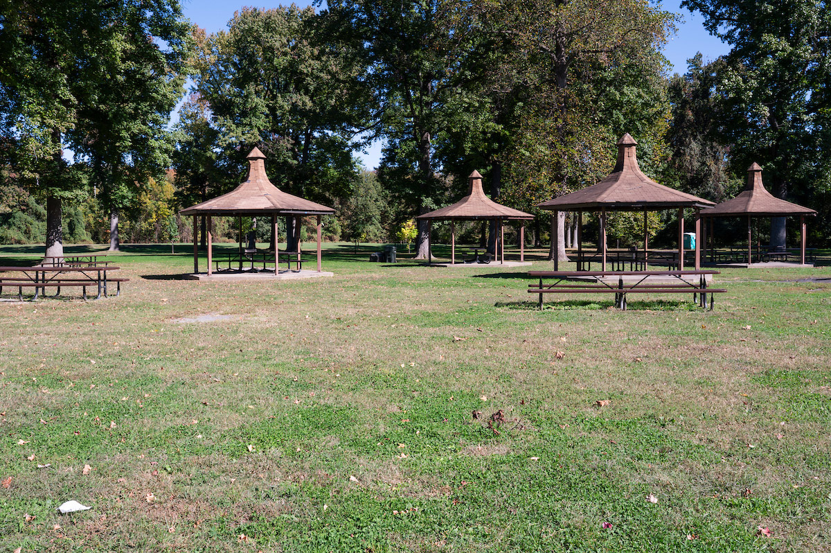 Five small picnic shelters in a field near some trees, each covering one picnic table. There are unsheltered picnic tables nearby.
