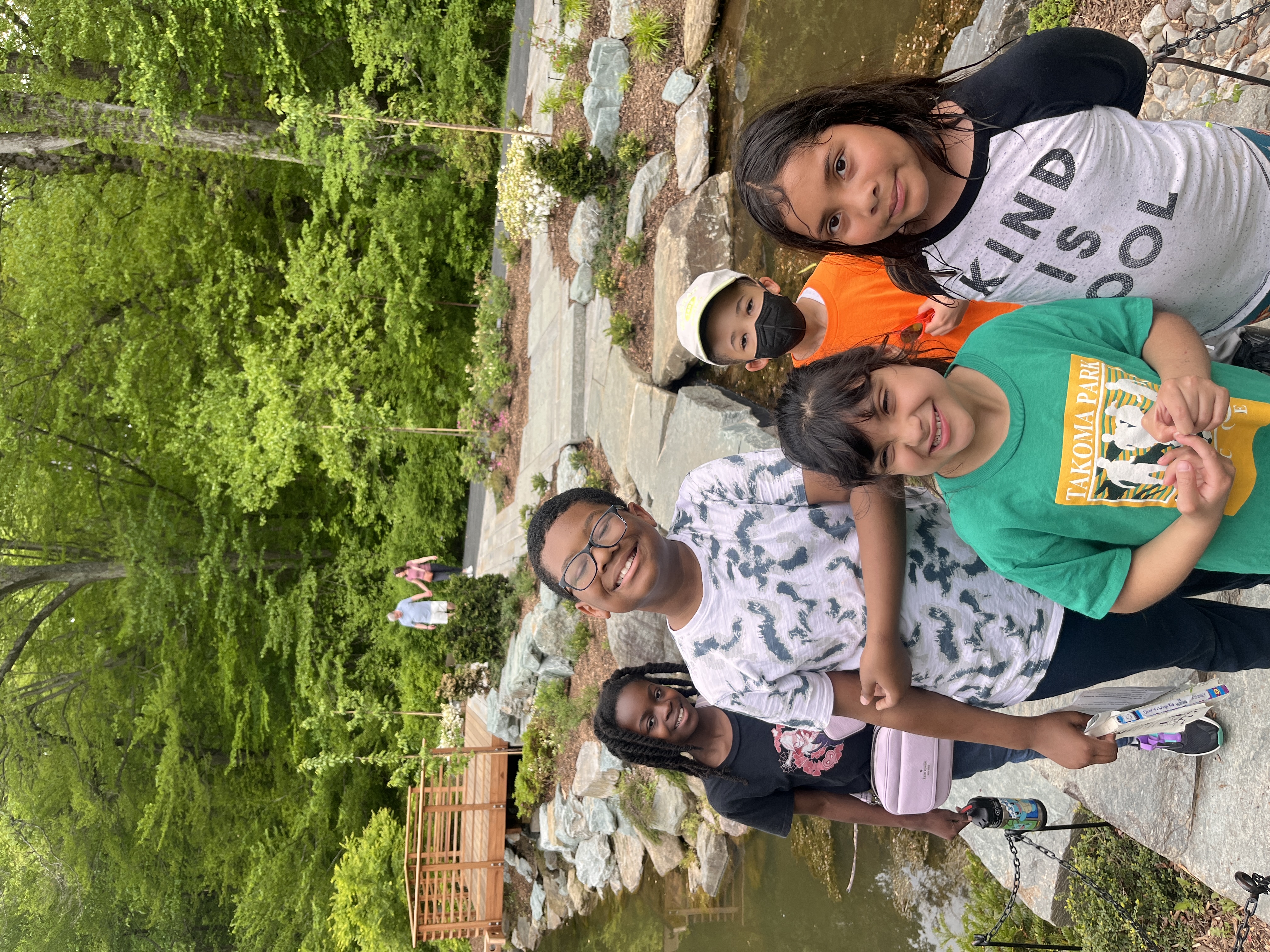 Five smiling kids standing by a rock bridge across a pond.