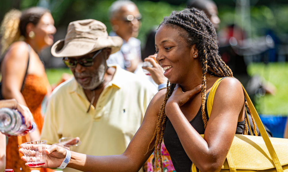 Person pouring a drink at an outdoor festival, with other people standing nearby on a sunny day.
