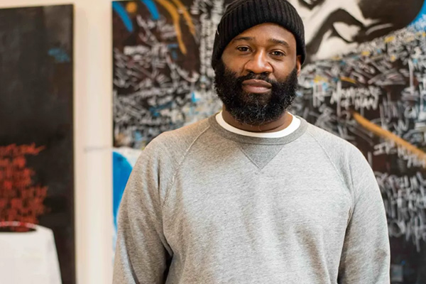 A man in a sweatshirt and knit cap standing in front of a wall where two large paintings are displayed.