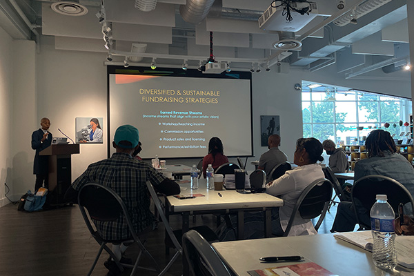 In a gallery, a speaker at a podium shares a slide presentation with a group of adults sitting at tables.