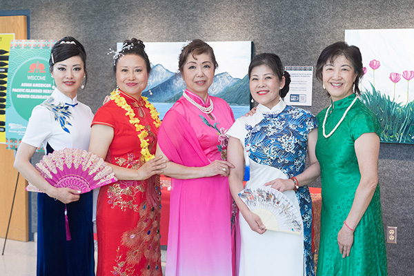 Five smiling women pose in an art gallery wearing traditional Chinese cheongsam dresses. Two are holding fans.