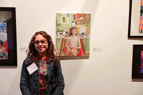 A smiling teen wearing a name tag poses next to a detailed self-portrait hanging on the wall in an art gallery.