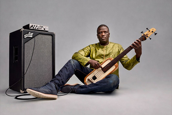 A man sits on the floor holding a very unique, oblong bass guitar that is plugged into an amp.