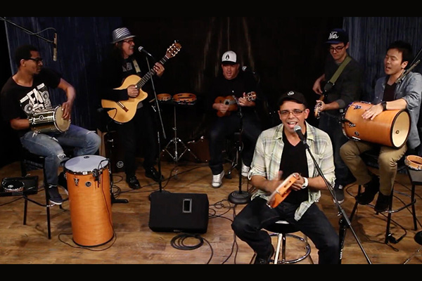 A six-piece band singing and playing various instruments, including hand-held drums, guitars, a ukulele, and a tambourine.
