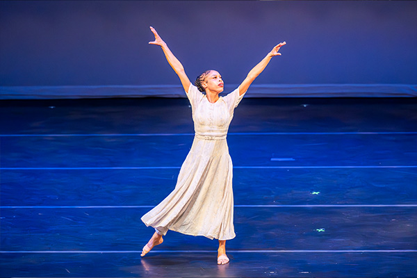 A woman in a long skirt dances barefoot on a stage with her arms raised.