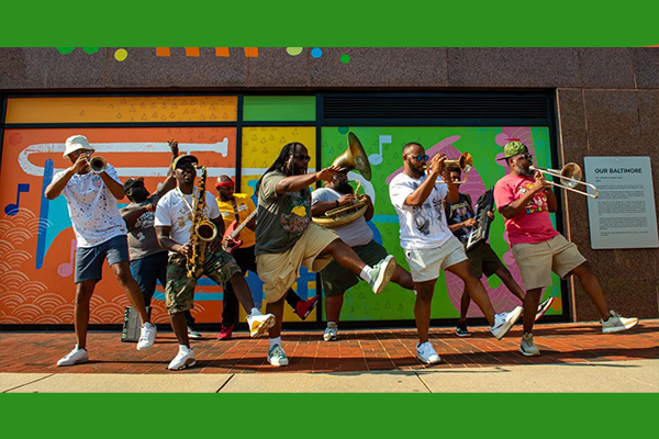 A nine-piece brass band plays their instruments while doing synchronized choreography on a sidewalk by a colorful storefront.