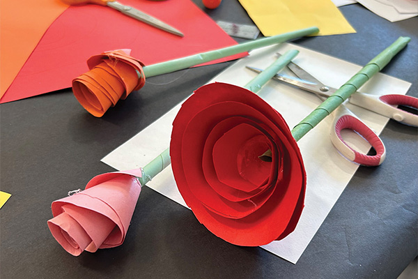 Three 3-D flowers made of colored paper lying on a table near scissors and paper.