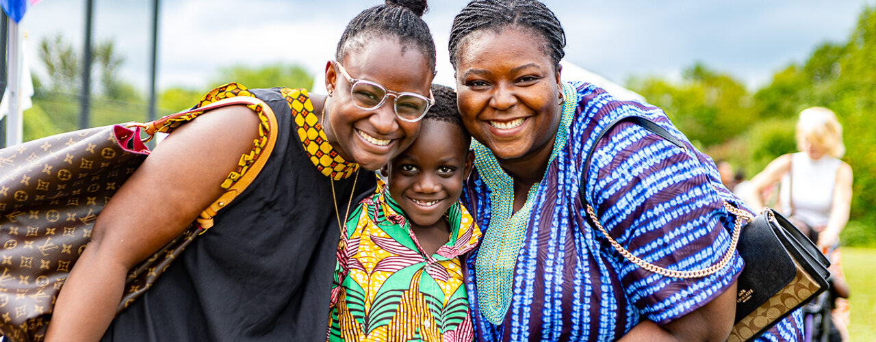 Three people pose closely together at an outdoor festival, wearing vibrant patterned clothing against a backdrop of grass, trees, and gathering attendees.