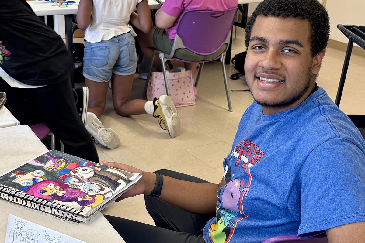 A young person sitting in a classroom holds a sketchbook containing a colorful drawing.