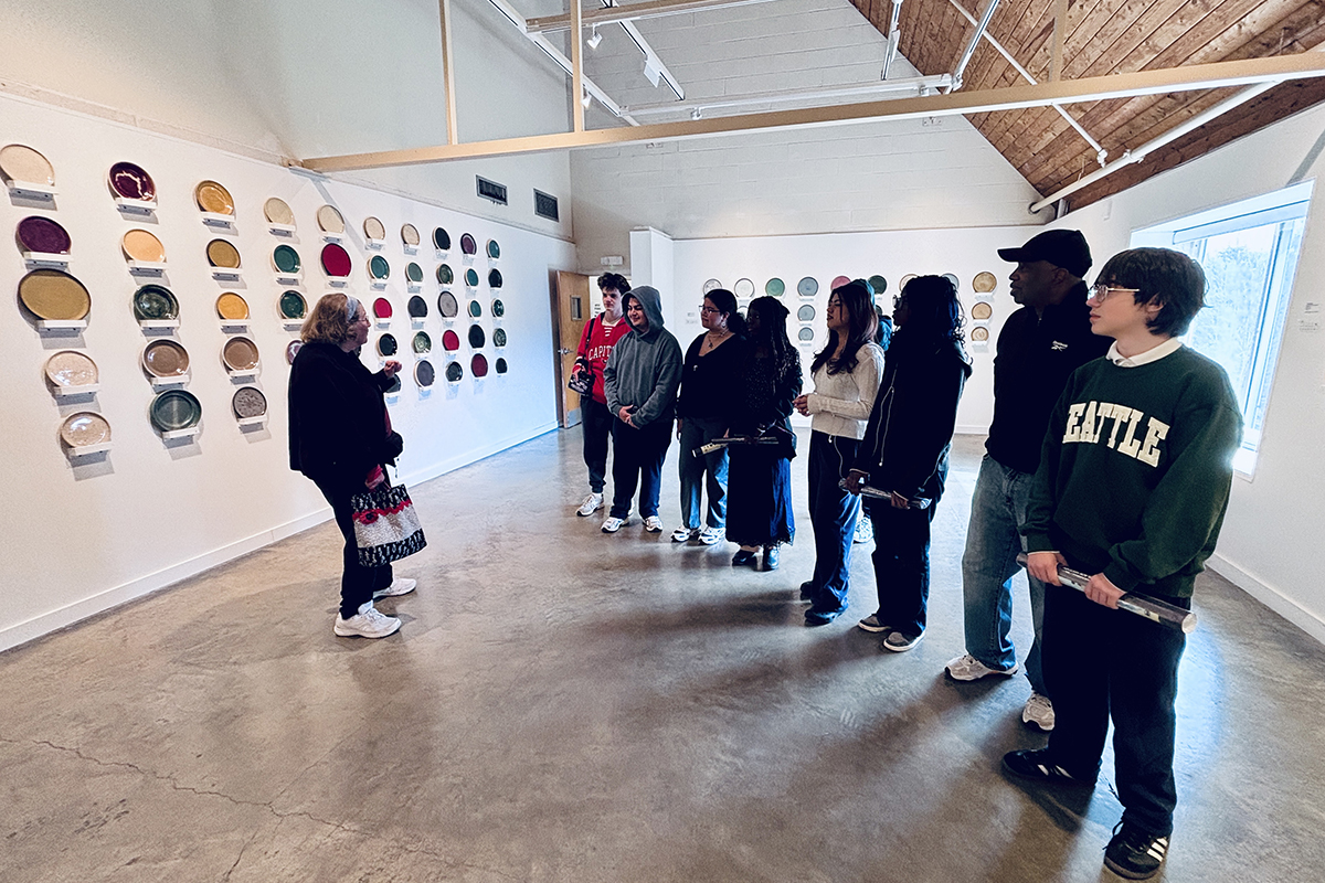 An artist stands in an art gallery, addressing a group of people. Dozens of ceramic plates in multiple shapes and colors hang on the walls.