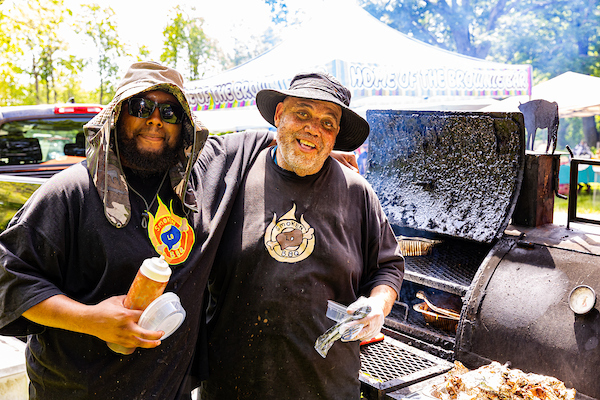 Two barbecue cooks pose beside a smoker grill at an outdoor community festival.