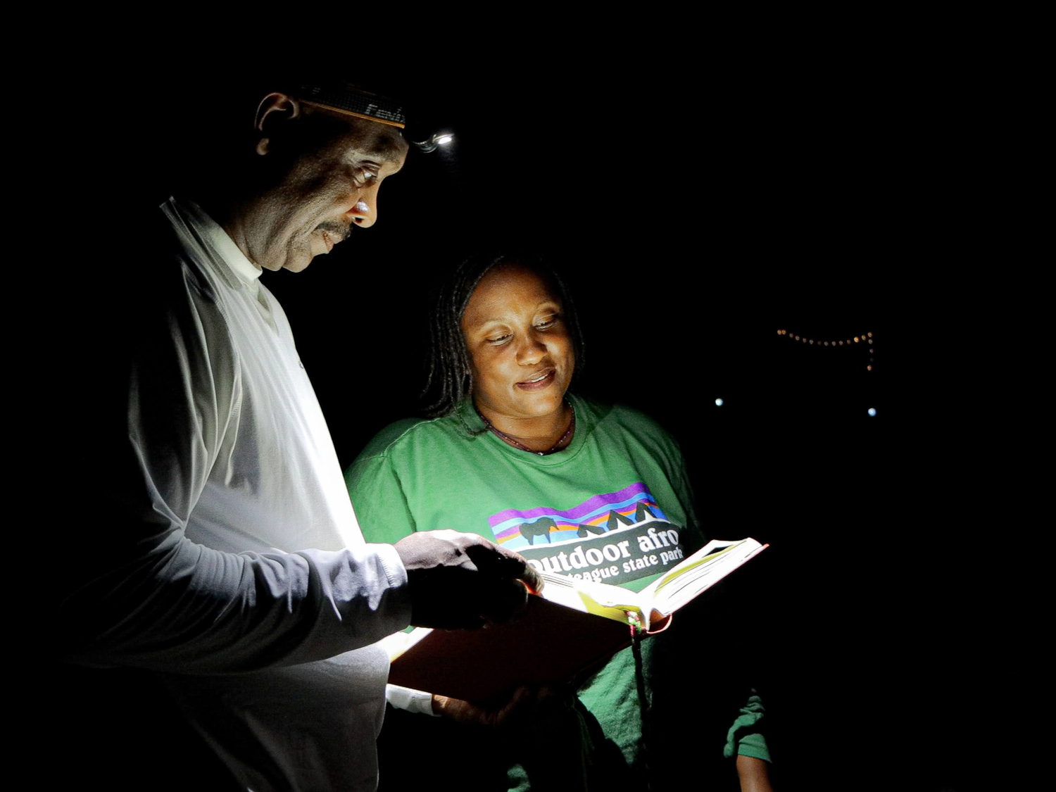 Two people read from an open book outdoors at night, illuminated by a bright light.
