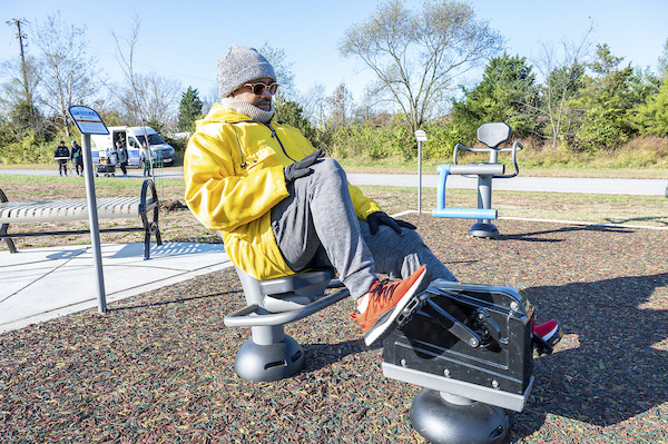 Person using a seated leg press machine at an outdoor fitness park, exercising on a rubberized surface with additional equipment in the background.