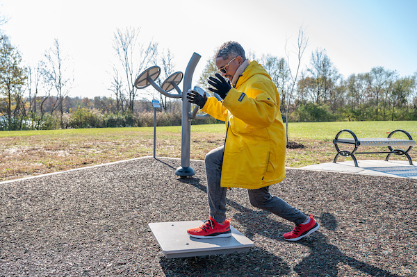 Person using a balance platform at an outdoor fitness park, performing a lunge exercise on a rubberized surface with exercise equipment nearby.