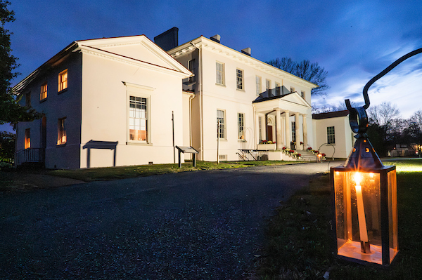 Historic manor house illuminated at dusk, with a lantern glowing beside the driveway during a candlelight tour event.
