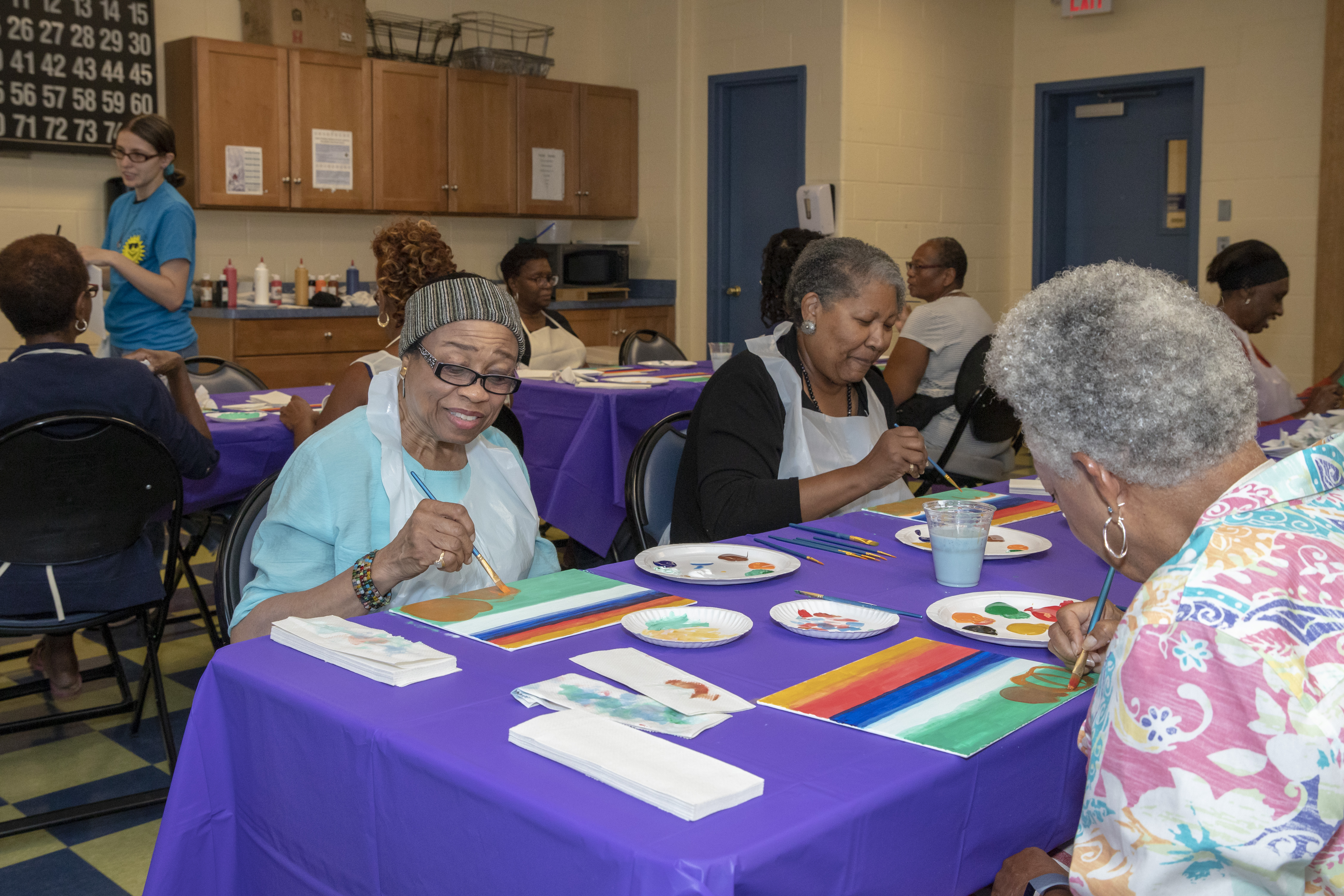 Seniors sitting around tables covered in purple tablecloths, painting colorful stripe designs on art canvases.