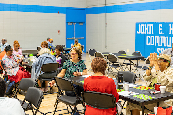 A large group of seniors seated at tables playing bingo and clapping inside a spacious community center gymnasium.