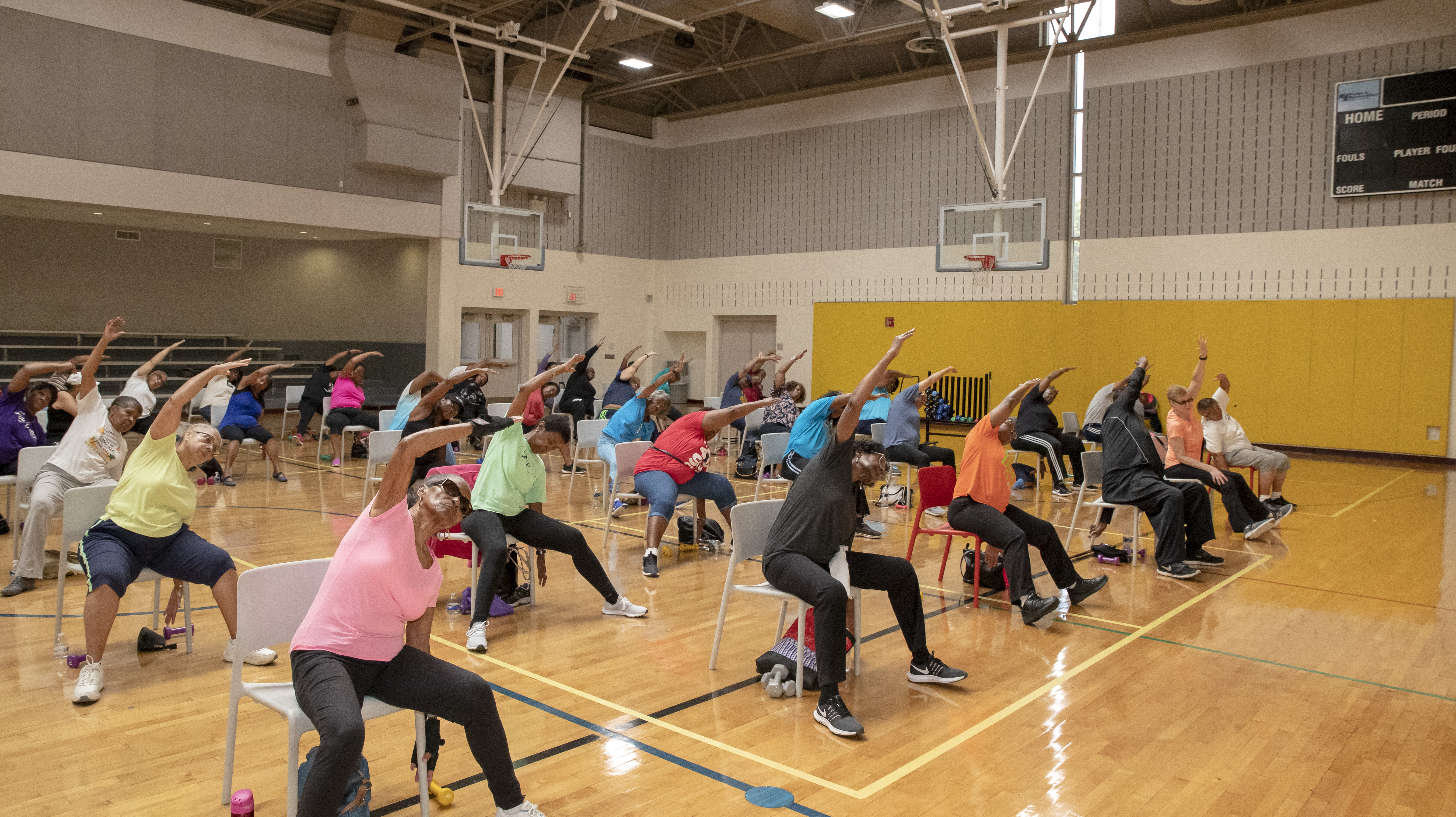 A large group of seniors performing side stretches while seated in chairs inside a gym.
