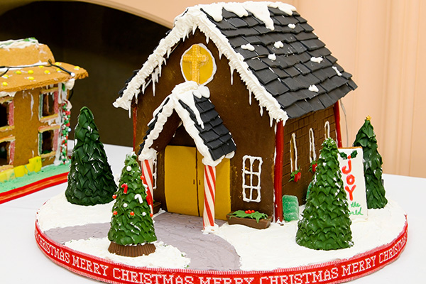 A decorated gingerbread house on a table, which appears to be a small church with candy cane porch pillars, icing "snow," and trees around it.