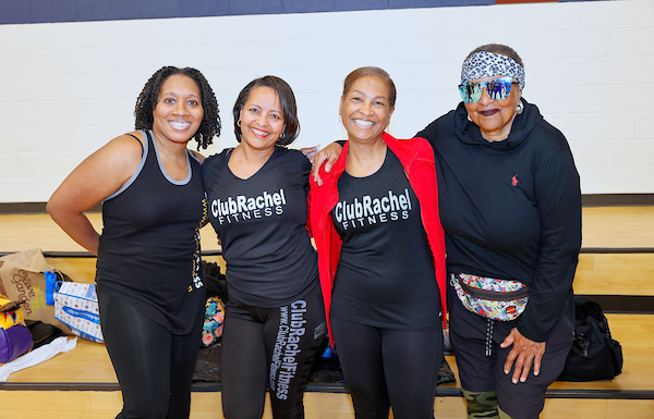 Four smiling women posing together inside a gym, three wearing matching "ClubRachel Fitness" shirts.