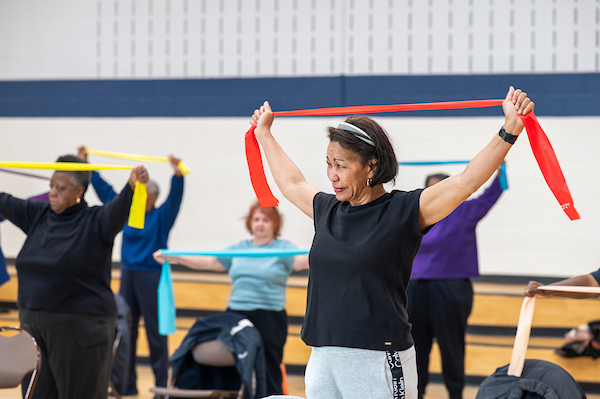 A group of active seniors stretching red, yellow, and blue resistance bands overhead during an indoor exercise class.