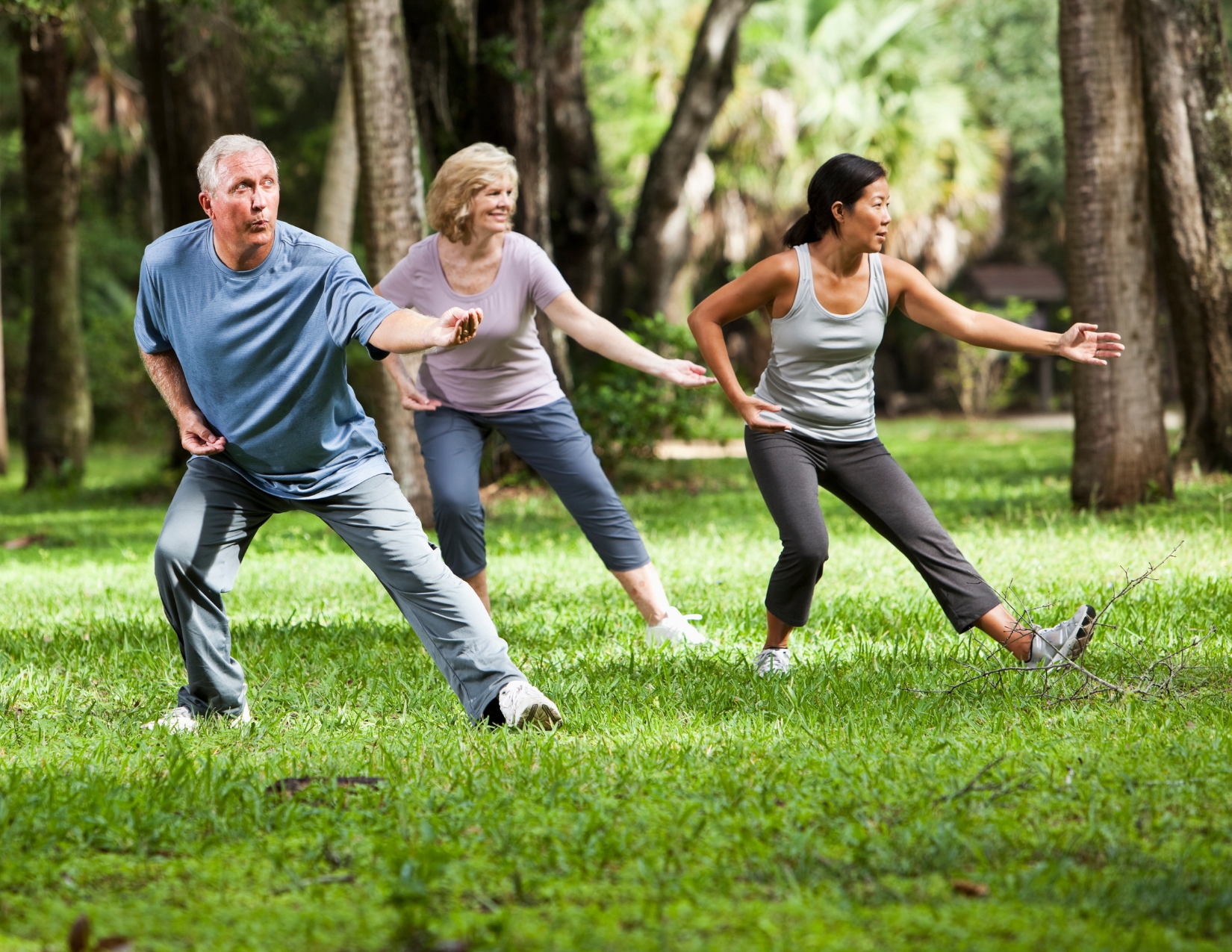 Three adults practicing low-impact Tai Chi stances together on a grassy lawn surrounded by trees.